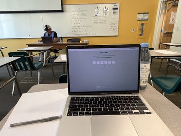 A photo of a MacBook laptop on a classroom desk with a blue login screen, with a person sitting at a desk working on a laptop in the background near a whiteboard covered in writing.