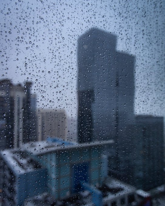 A view through a rain-covered window showing a blurred cityscape with tall skyscrapers in the background under overcast skies.