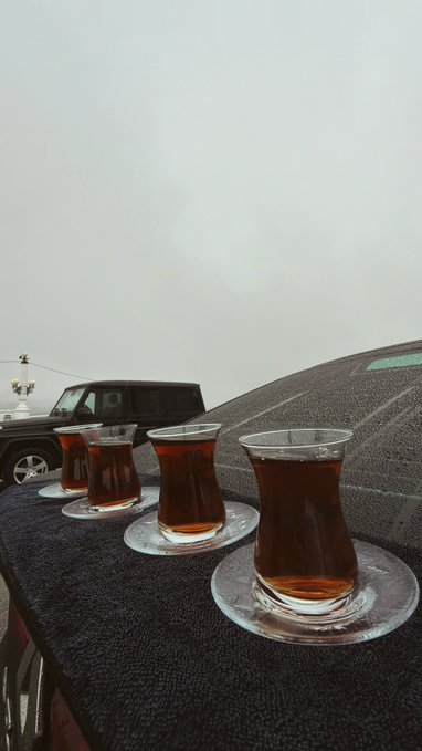 Four glasses of dark tea, likely chai, in traditional curved glassware sit on saucers arranged on a black towel, likely placed on a car dashboard against a foggy, overcast background.