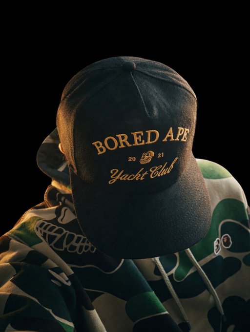 The image features a person wearing a black baseball cap with gold lettering that reads "Bored Ape Yacht Club". They are also wearing a green and white patterned hoodie, which complements their hat. The background of the photo is dark, making the colors of the hat stand out even more.