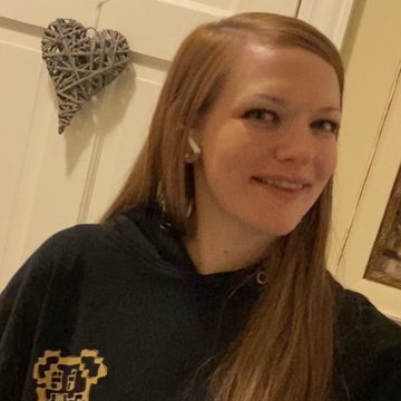 A selfie of a smiling woman with long reddish-brown hair wearing a dark hoodie and a wireless earbud, with a woven heart decoration hanging on a white paneled wall behind her.