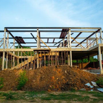 A house under construction with a wooden frame, surrounded by dirt piles and an excavator.