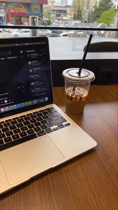 The image shows a laptop computer sitting on top of a wooden table with an open coffee cup next to it. The scene appears to be set in a cafe or office environment, as there are multiple cars visible outside the window. A person can also be seen in the background, possibly working or enjoying their time at the establishment.