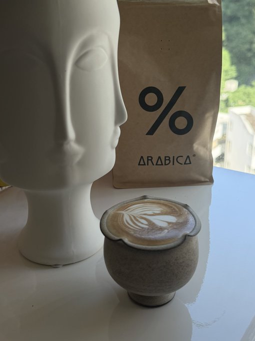 A latte with leaf latte art served in a speckled ceramic cup next to a white sculpted head and a brown bag of '% ARABICA' coffee.