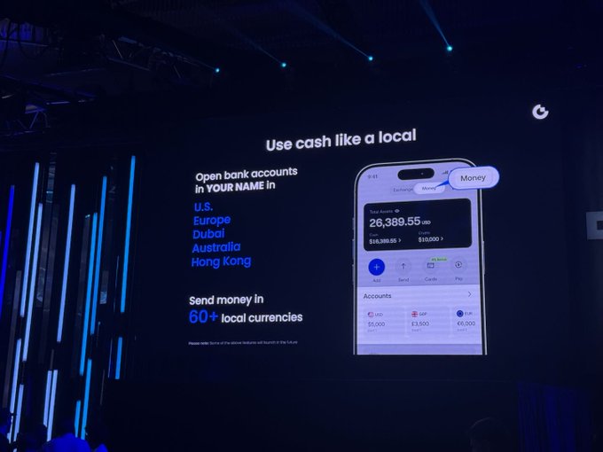A dimly lit presentation stage showing a large screen displaying a mobile banking app with the headline 'Use cash like a local'. The screen advertises opening bank accounts in 'U.S., Europe, Dubai, Australia, Hong Kong' and sending money in '60+ local currencies', with a user balance of 26,389.55 USD shown.