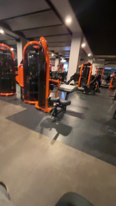 A dimly lit indoor gym filled with orange and black strength training equipment, including cable machines and benches, on a rubberized floor.