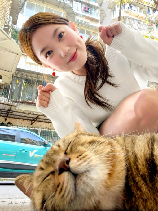 A selfie-style photo of a smiling young woman making fists, with a tabby cat in the foreground resting its eyes, set against a backdrop of a building and a teal taxi.