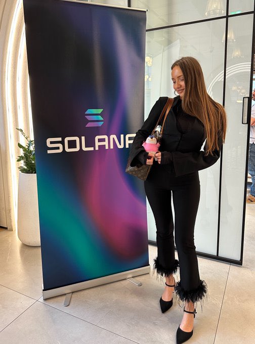 A woman in a black outfit with feathered boots poses next to a Solana-branded banner, holding a pink plush toy.