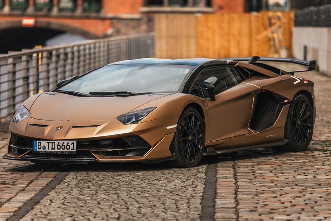 A matte bronze Lamborghini Aventador SVJ with a Berlin license plate reading "B TD 6661" sits parked on cobblestones, embodying the sleek innovation and ambitious performance that ties the Italian supercar brand to its partnership with Solana at the German summit. The car's aggressive aerodynamic design and commanding presence visually reinforce the post's message of shared values between cutting-edge automotive engineering and forward-thinking blockchain technology.