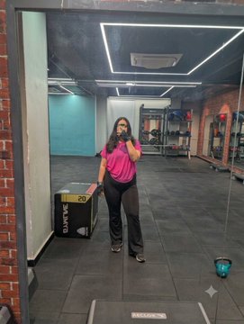 A woman in a pink top takes a mirror selfie inside a gym with equipment racks and an air conditioning unit visible.