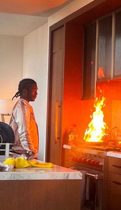 A dramatic photo of a person with dreadlocks standing calmly in a kitchen while large flames erupt from the stovetop.