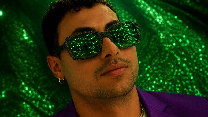 A man wearing sparkly green sunglasses and a purple blazer poses against a shimmering green background, projecting a confident, stylish vibe that mirrors the celebratory tone of the post about personal and financial milestones in the Web3 creator space.