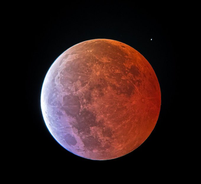 A photograph of a reddish-orange moon, likely during a lunar eclipse, set against a black sky with a small bright point of light nearby.