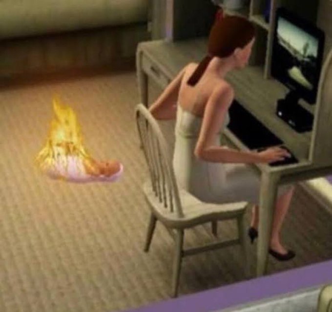 A Sims character in a towel sits at a desk using a computer while a small fire burns on the floor nearby.