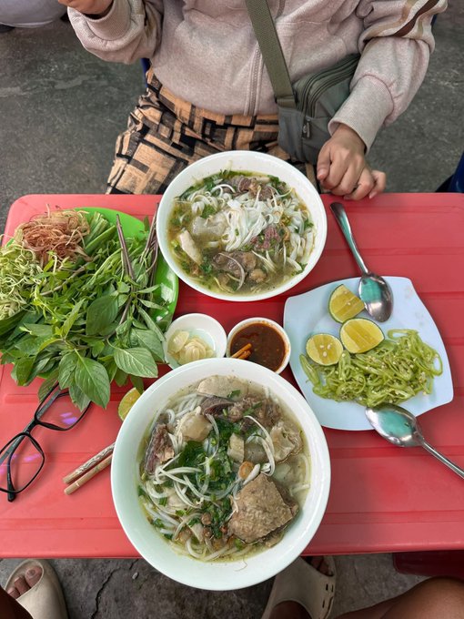 Two bowls of Vietnamese pho with meat and herbs sit on a red plastic table, accompanied by fresh herbs, bean sprouts, lime wedges, chili sauce, and a child's hands visible at the top.