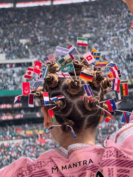 A fan at a large stadium sports numerous country flags—including Iran, Canada, Saudi Arabia, Brazil, Germany, and the Netherlands—adorned in her braided hair, wearing a jersey with "MANTRA" printed on the back, symbolizing global unity and diversity that mirrors the post's message of "48 nations, one purpose-built EVM L1 for RWAs."