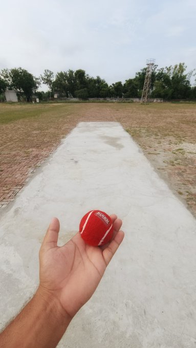 A hand grips a red cricket ball marked "DSC" on an outdoor pitch, capturing a fresh "good green morning" with a pitch set for a new innings.