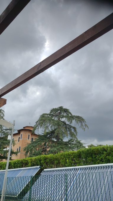 A cloudy, overcast sky viewed from beneath a wooden pergola structure, with a brick building, a large green tree, hedges, and a metal fence visible in the lower portion of the frame.