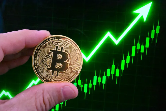 A hand holds a physical Bitcoin token in front of a green upward-trending price chart.