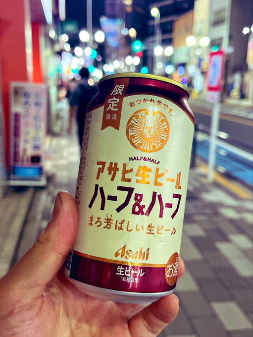The image features a person holding up an open can of beer, which has Japanese writing on it. The text "半分だけ、おつかれ生です" translates to "Half-lager". This suggests that the person is showing off their half-lager drink choice or possibly discussing its taste with someone else who might be interested in trying it out.

The background of the image shows a city street, indicating that this beer may have been purchased from a local store or market. The presence of other people and buildings suggests that they are likely in an urban setting.