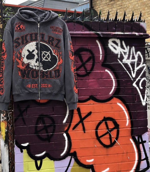 A dark grey zip-up hoodie with skull graphics and 'SKULLRZ WORLD' text, along with 'EST. 2022', displayed against a graffiti-covered wall.