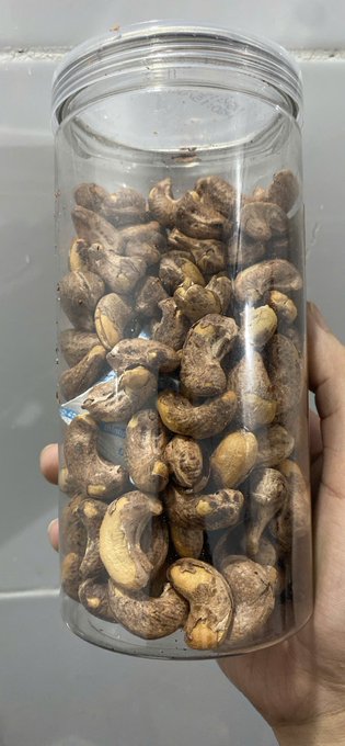 A hand holds a clear plastic container filled with raw cashew nuts still in their shells, with a small packet visible among them.