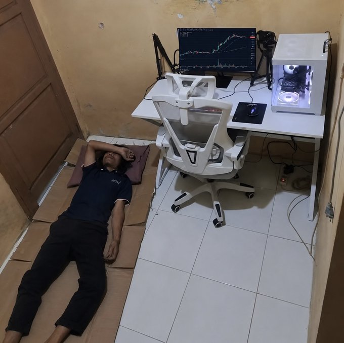 A man lies on cardboard mats on the floor in a small room, seemingly asleep or resting, positioned right next to a large white desk setup with a gaming PC, monitor displaying a trading chart, and a white gaming chair.