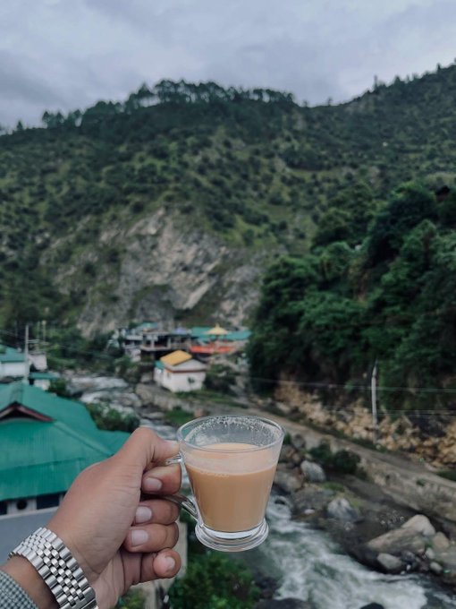 A hand holding a glass cup of tea or chai in front of a scenic mountain view with buildings and a river below.