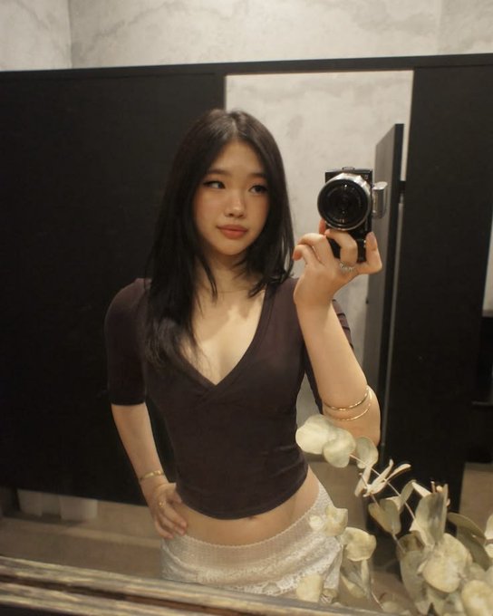 A mirror selfie of a young woman with long dark hair wearing a brown v-neck top and a light-colored skirt, holding a camera with eucalyptus leaves visible in the foreground.