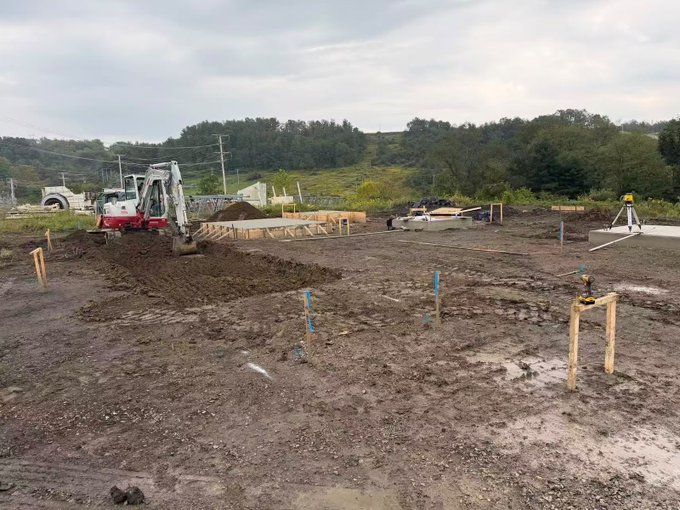 Construction site photo showing an excavator digging foundations for a building, with wooden forms set up for a concrete slab, surrounded by dirt ground and trees in the background.