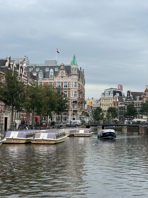 A scenic view of Amsterdam canal boats docked along the water with historic buildings and a green-domed tower in the background under an overcast sky.