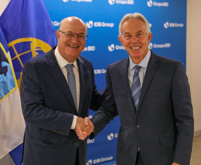 The image shows two men shaking hands, both wearing suits and ties. They are standing next to a blue wall with a flag displayed behind them. The background of the image features an IDB Invest blog that discusses the future of government modernization through the lens of smart mining in Latin America. This particular post is about blockchain technology and digital traceability, which could be related to the men's discussion on modernizing governments or improving transparency and efficiency within public services.

The main focus of the image seems to be the handshake between the two men, as they are positioned close together with their hands meeting in a firm grip. The presence of the flag behind them suggests that this event might have some significance or connection to the IDB Invest blog's topic.
