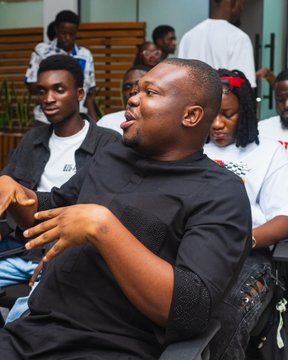 A man in a black polo shirt sits with his hands gesturing outward while speaking, surrounded by several people seated behind him in what appears to be an indoor gathering.