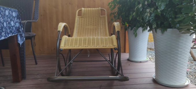A yellow woven rocking chair sits on a wooden porch, positioned between a table with a blue floral cloth and two white planters containing green plants.