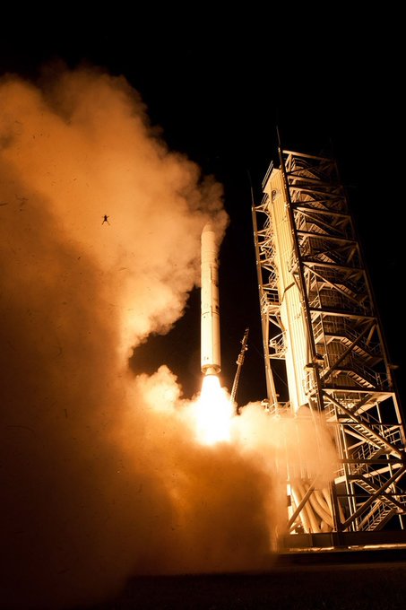 A nighttime photograph captures a rocket launching from a tower, with bright flames and billowing clouds of smoke illuminating the scene.