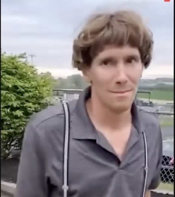 A photo of a man with distinctive bowl-cut hair and a slight smile, wearing a dark collared shirt, standing outdoors near a chain-link fence with greenery in the background.