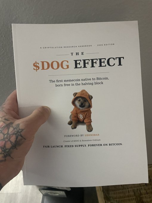 A tattooed hand holds a printed booklet titled 'THE $DOG EFFECT' which describes itself as 'The first memecoin native to Bitcoin, born free in the halving block,' featuring a Shiba Inu dog wearing an orange Bitcoin hoodie.
