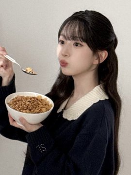 A young woman with long dark hair wearing a navy sweater with a white collar holds a bowl of cereal and a spoon, looking at the camera with puffed cheeks.