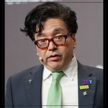 A man wearing glasses, a dark suit, and a green tie is shown, with a small cross pin and a "BMNR" ticker badge visible on his lapel. The image relates to the post as it likely depicts Tom Lee, the figure associated with Bitmine (ticker: BMNR) who repurchased 5.5 million shares amid reduced ETH acquisitions of 7,430 tokens.
