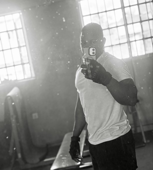 A black-and-white photo of a muscular, bald man taking a mirror selfie in a gym, wearing a white t-shirt and fingerless gloves, with light streaming through large windows behind him.