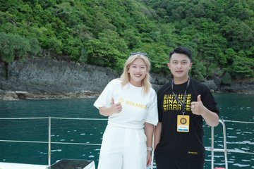 A casual photo of two young people on a boat with turquoise water and green cliffs in the background; the woman wears a white 'BOSS' t-shirt and the man wears a black graphic tee, both giving thumbs up.