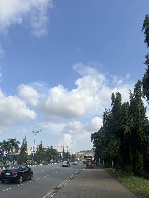 A street scene showing a road with cars, trees lining the sidewalk, and a blue sky with scattered clouds.