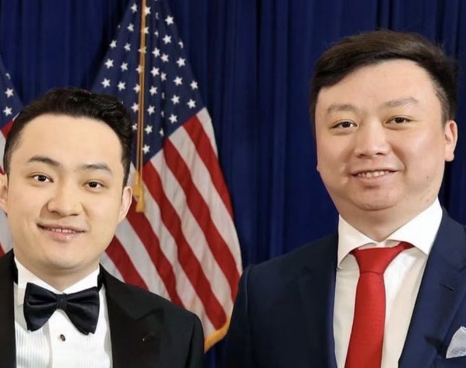 Two men in formal attire—one in a black tuxedo with bow tie, the other in a dark suit with red tie—pose in front of American flags, appearing to represent HTX figures Justin Sun and HTX_Molly referenced in the post about loyalty and compensation.