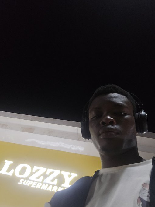 A nighttime selfie of a young man wearing over-ear headphones and a white t-shirt, standing in front of a brightly lit yellow sign that reads 'LOZZY SUPERMARKET'.