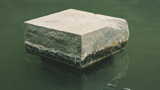 A weathered rectangular stone block partially submerged in green water with a dark algae/waterline stain around its base.