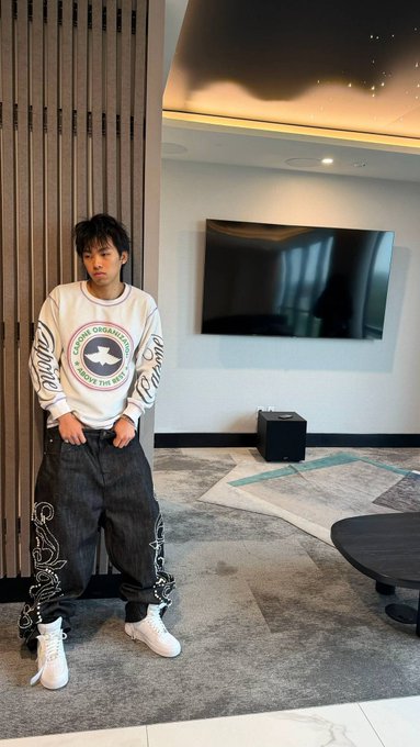 A young man poses indoors wearing a white graphic sweatshirt and dark patterned pants, standing near a wall-mounted TV in a modern room.