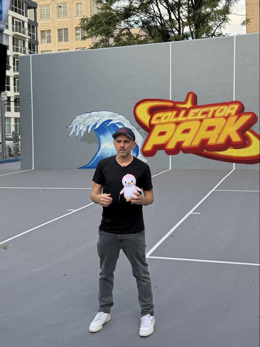 A man in a black t-shirt featuring a pink penguin, jeans, and a cap stands on a court with a colorful 'Collector Park' mural behind him.