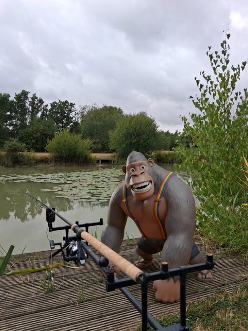 A humorous, surreal scene of a grey gorilla statue wearing rainbow suspenders, kneeling next to a fishing rod on a wooden dock by a lake.