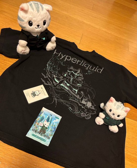 A black t-shirt with 'Hyperliquid' graphic design laid on a wooden floor, accompanied by two white cat plush toys wearing dark outfits, a small card, and a 'GENESIS' candy wrapper.