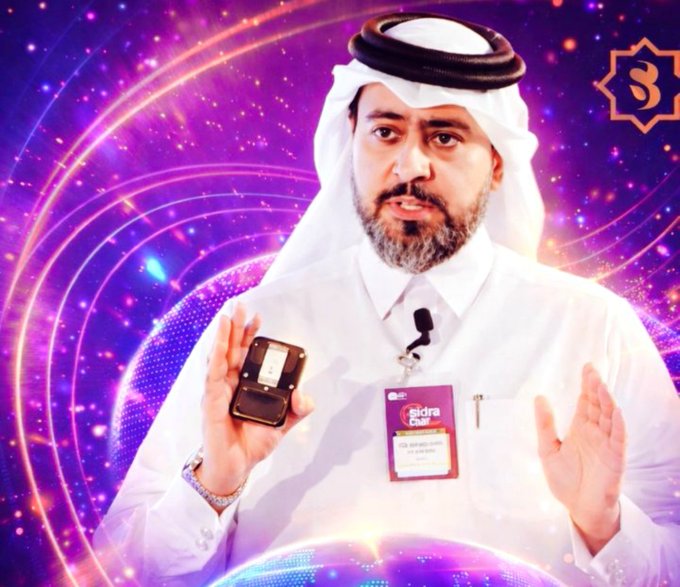 A man wearing traditional Middle Eastern attire (white thobe and ghutra) holds a small black device while speaking, with a name badge reading 'Sidra Qatar' and a purple cosmic background behind him.